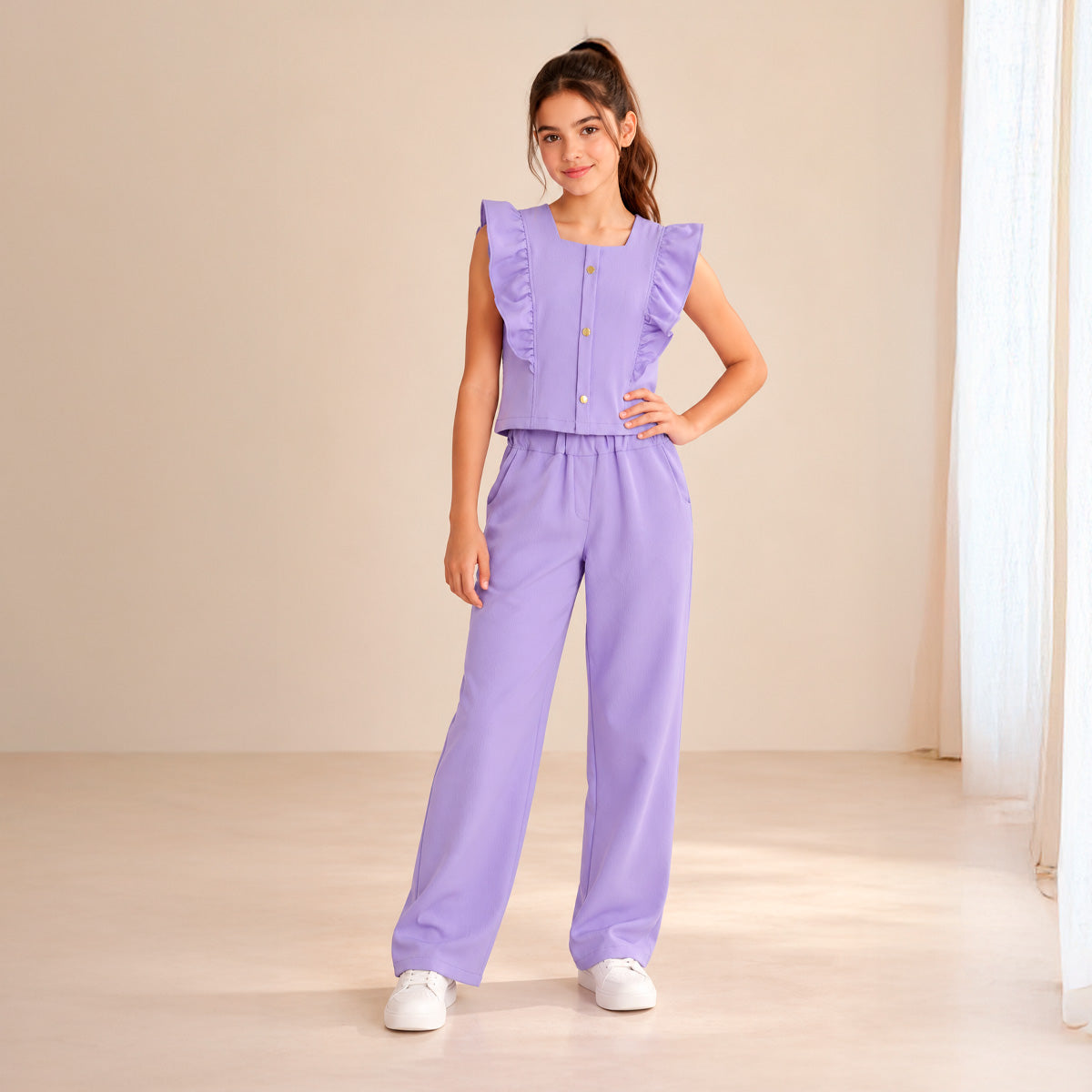 Niña con conjunto lila de blusa sin mangas con olanes y botones al frente, pantalón largo y tenis blancos.