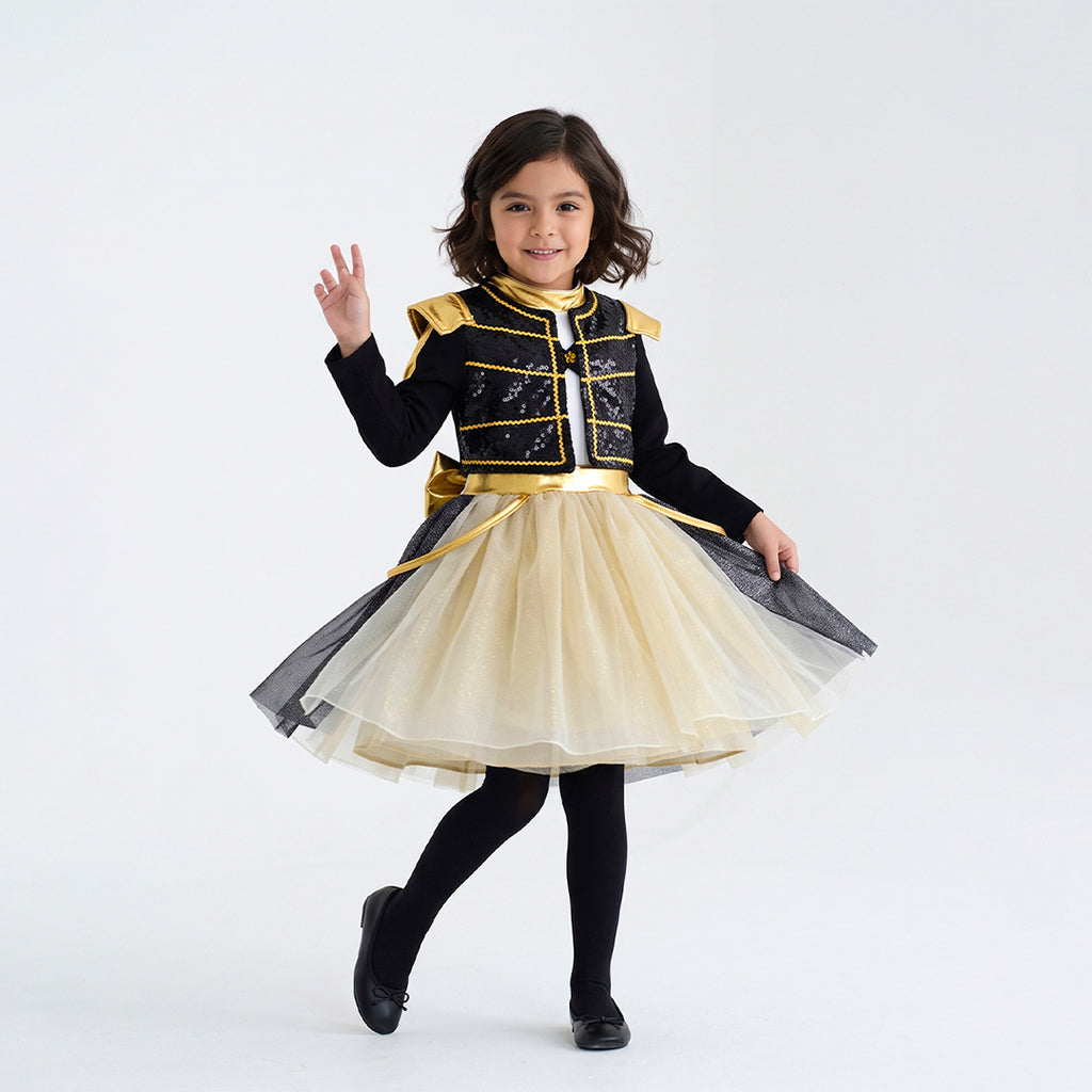 Niña modela conjunto infantil negro con detalles dorados y falda de tul color crema, combinado con medias y botas negras.