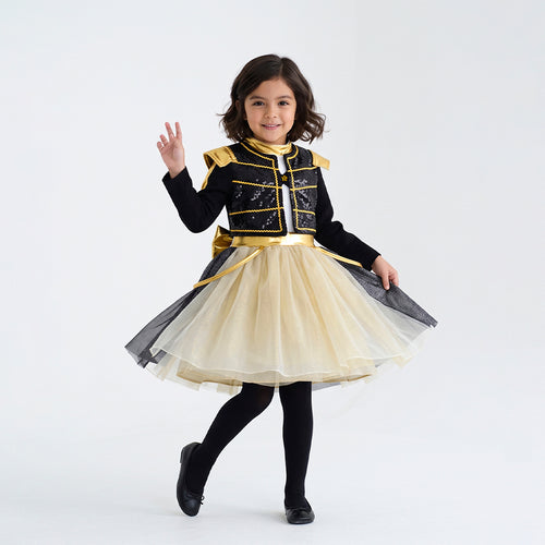 Niña modela conjunto infantil negro con detalles dorados y falda de tul color crema, combinado con medias y botas negras.