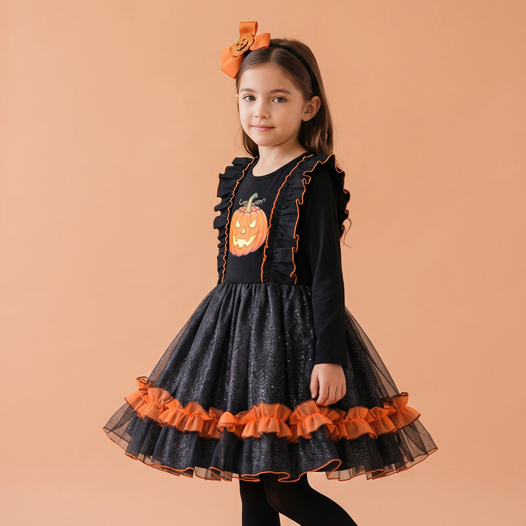 Niña modela vestido negro de Halloween con estampado de calabaza, falda de tul con olán naranja y diadema con moño naranja, sobre fondo durazno.