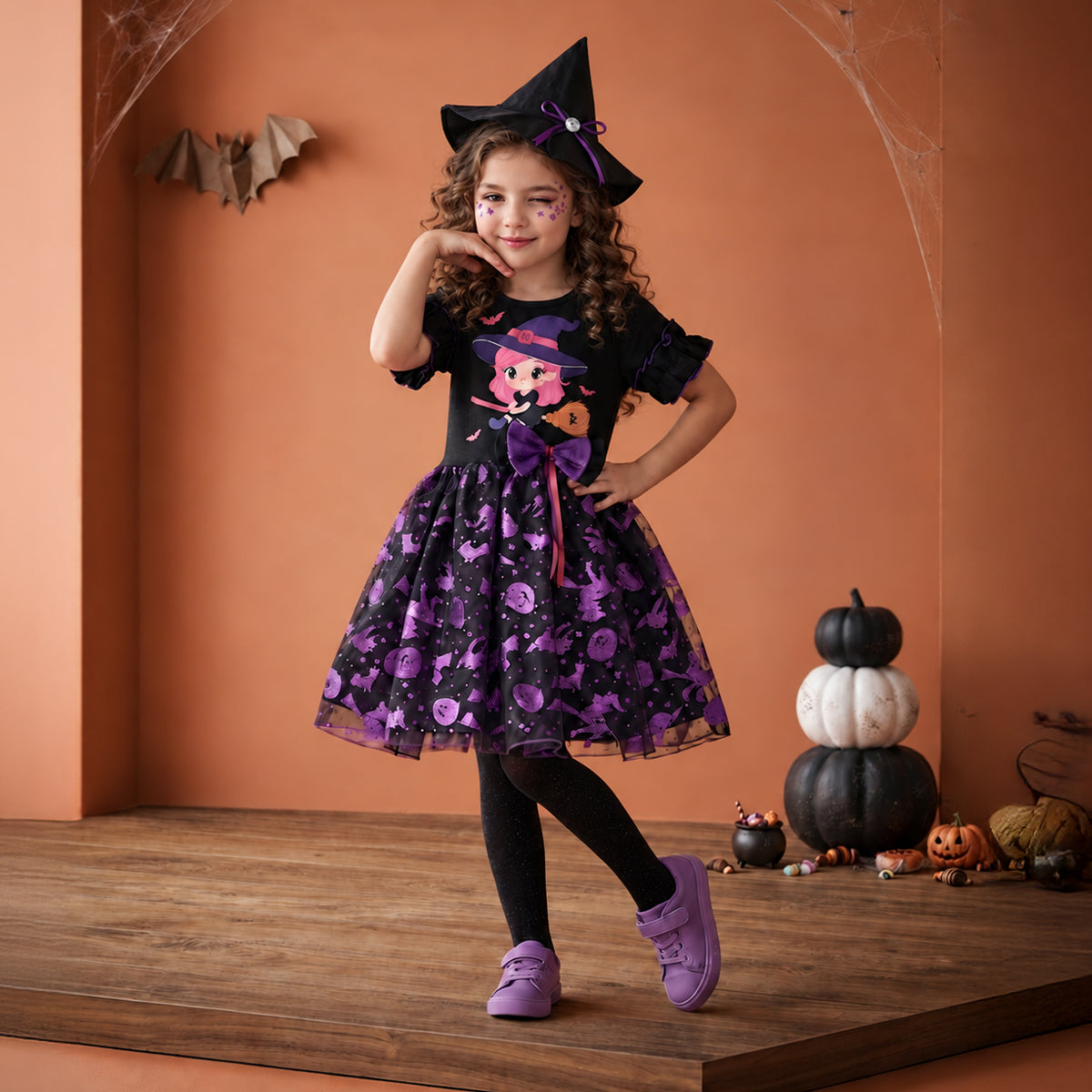 Niña con vestido de brujita negro y morado, sombrero puntiagudo, medias negras y zapatos morados, junto a calabazas.
