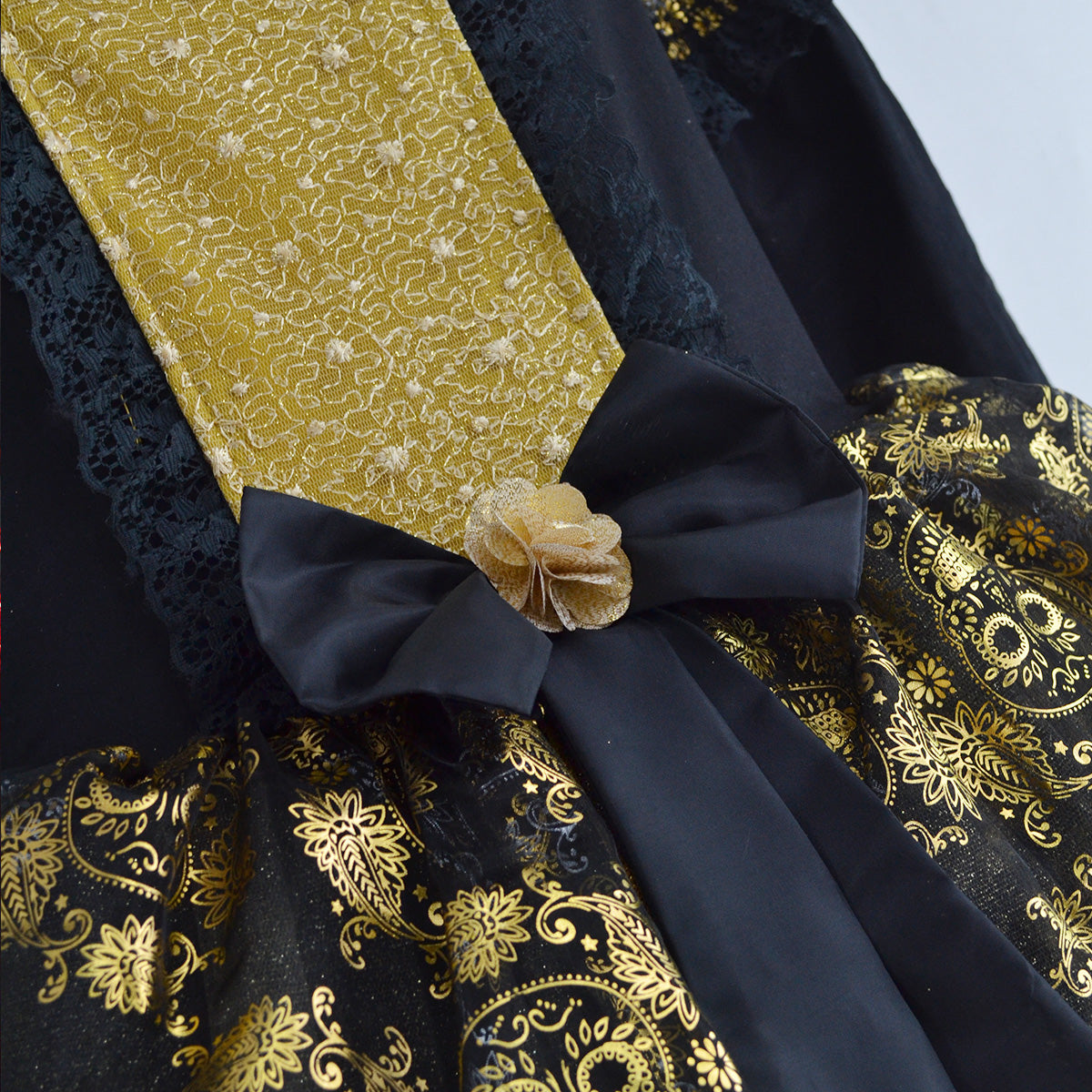Detalle del vestido negro con panel dorado, moño negro y flor dorada en la cintura, sobre tela estampada con calaveras y flores.