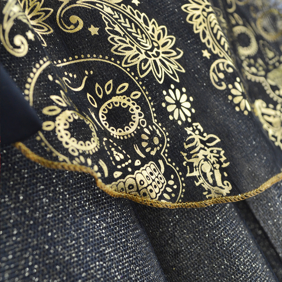 Detalle de tela negra con estampado dorado de calaveras y flores, ribete dorado y acabado brillante.
