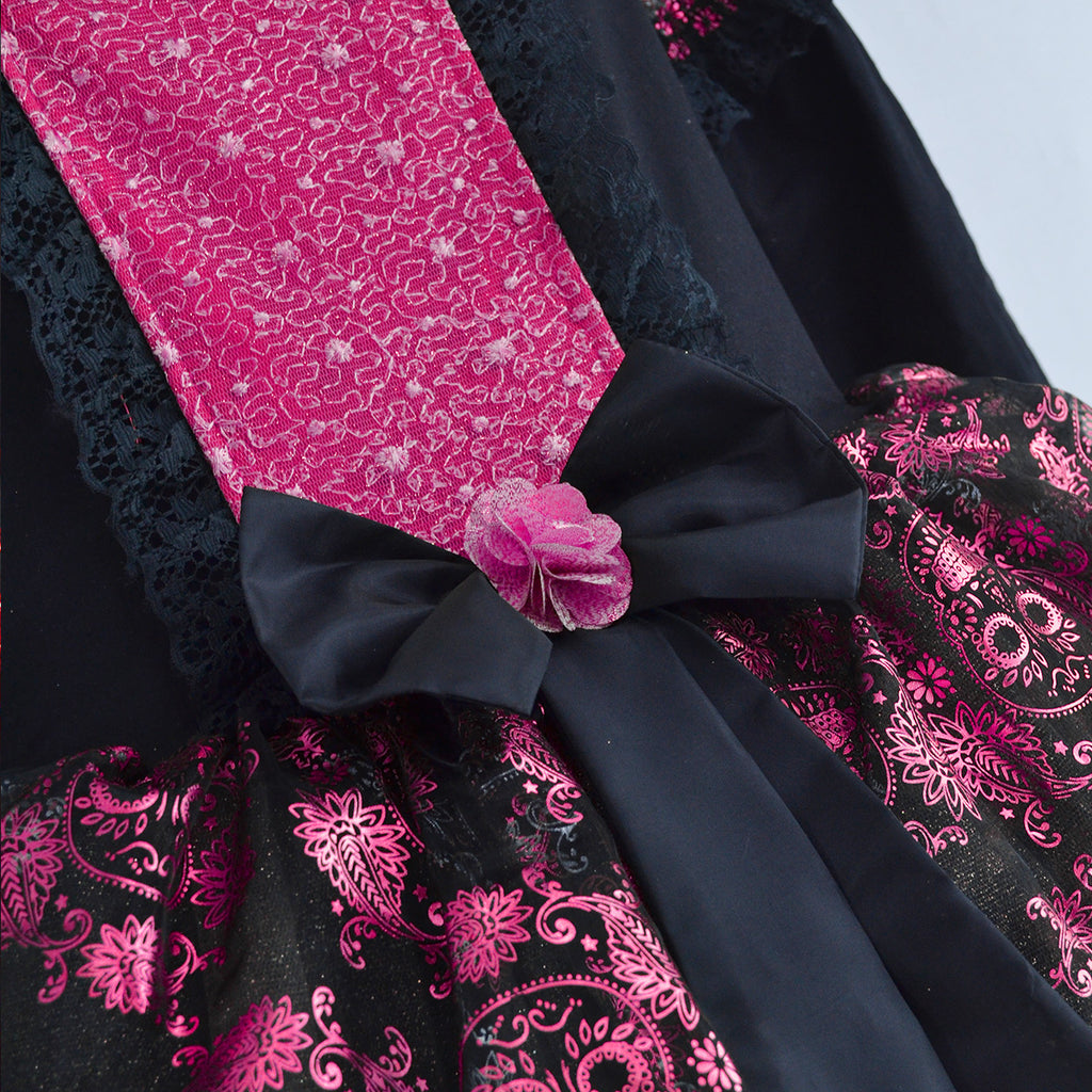 Detalle del vestido negro con panel fucsia, moño negro y flor rosa en la cintura, sobre tela estampada con calaveras y flores.